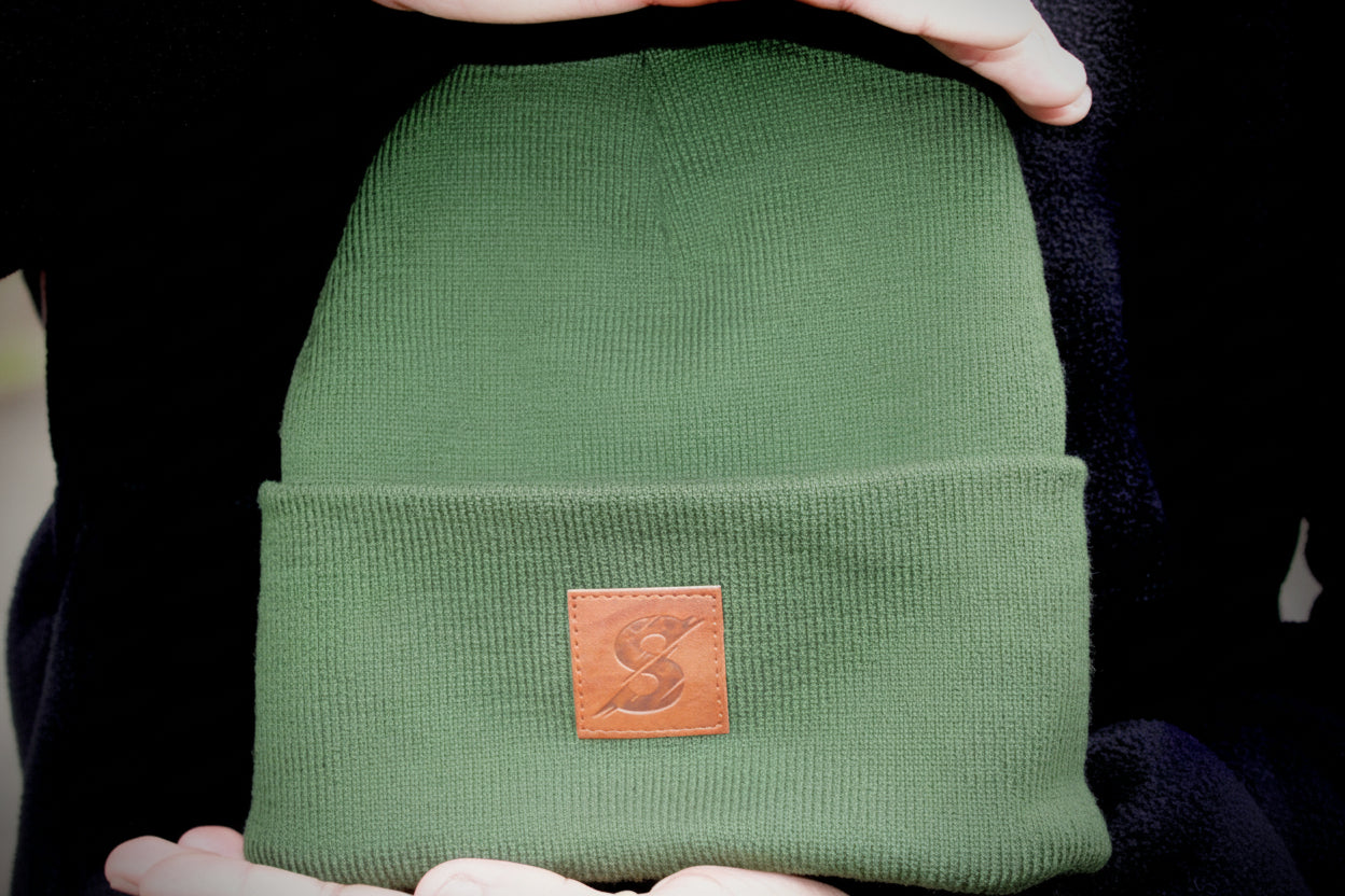 Green beanie with a brown leather patch held by hands against a black background