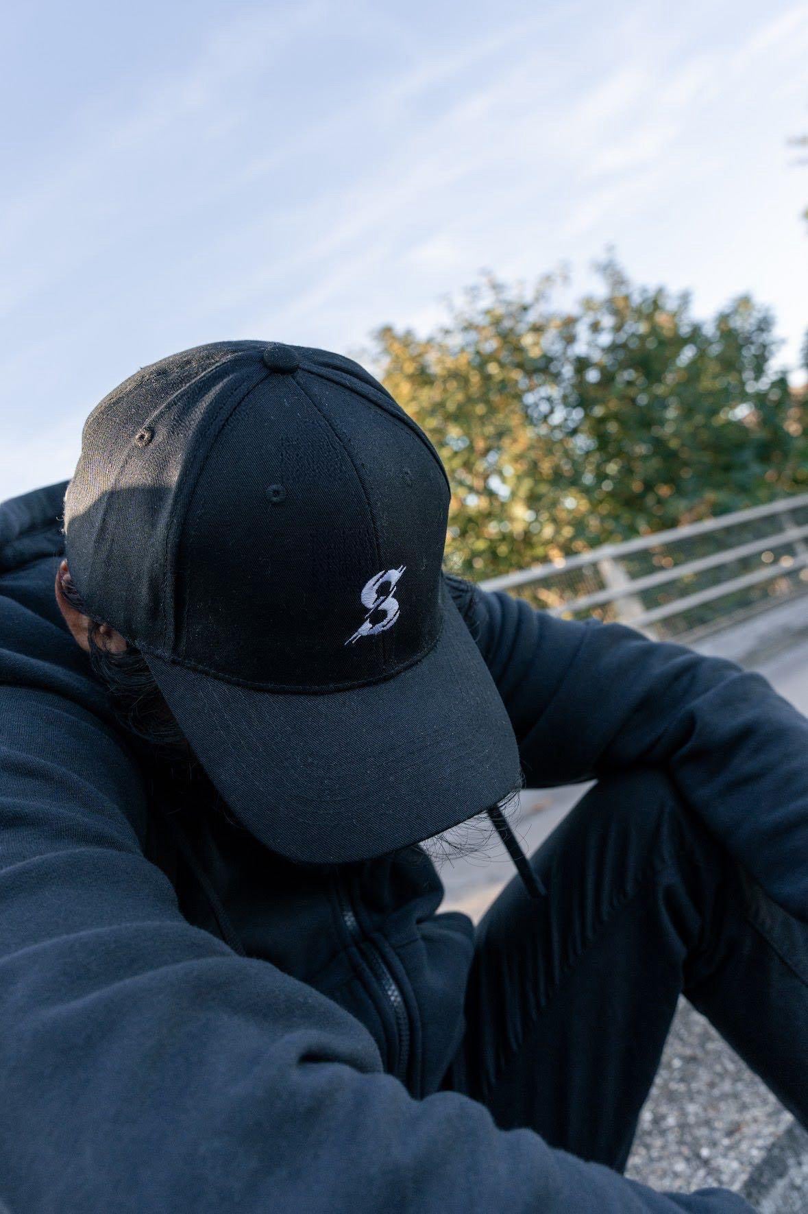 Person wearing a black cap with a logo, sitting outdoors with trees in the background