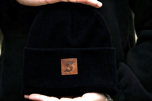 Black beanie with a brown leather patch held in hands against a dark background