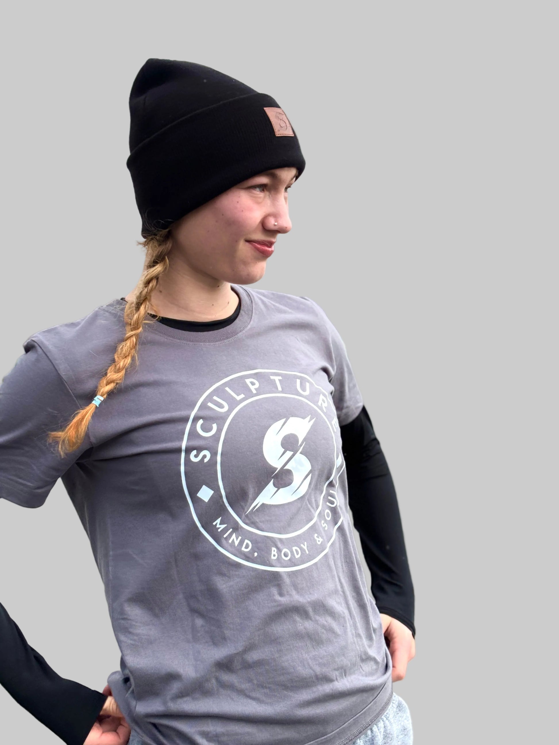 A woman wearing a black beanie and a gray graphic t-shirt with a white circular logo that reads 'SCULPTURE MIND. BODY.