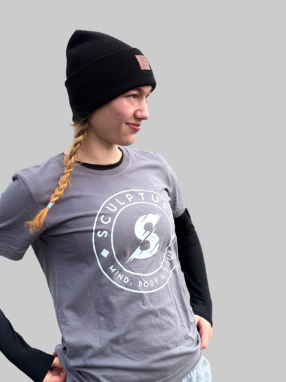 A woman wearing a black beanie and a gray graphic t-shirt with a white circular logo that reads 'SCULPTURE MIND. BODY.