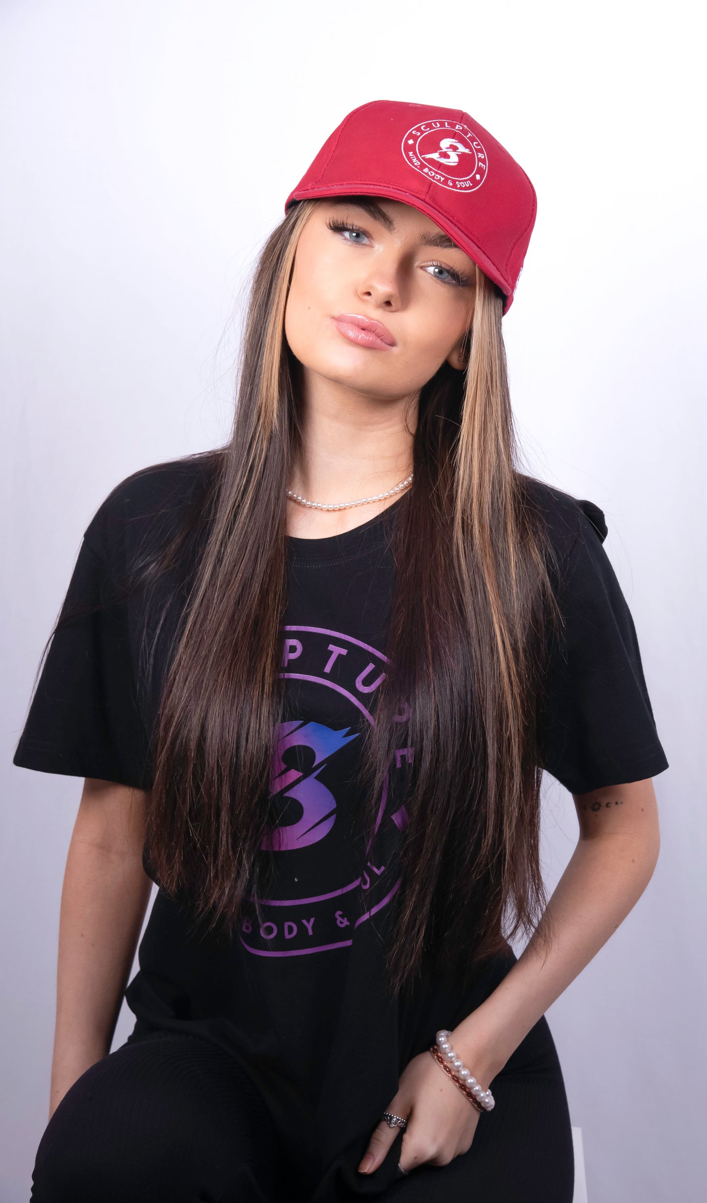 A woman wearing a red baseball cap with a white circular logo, a black graphic t-shirt, and black pants, posed against a light gray studio background.