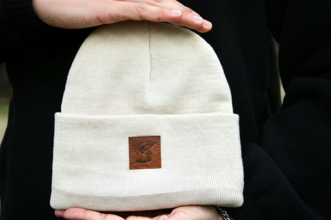 Beige beanie held by a person with a brown leather patch, on a blurred background.